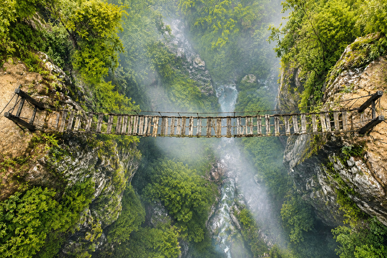 Vu d’en haut, un pont suspendu de planches de bois traverse un canyon couvert de végétation, au-dessus d’une rivière noyée de brume.